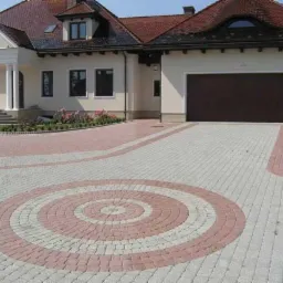 Elegancki dom z jasną elewacją i ciemnym dachem, ozdobiony kolumnami przy wejściu, z podjazdem wyłożonym kostką brukową w geometryczny wzór z okręgami w odcieniach szarości i różu.