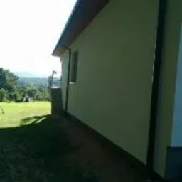 Świeżo pomalowana, jasnozielona ściana domu jednorodzinnego z widoczną rynną, małym oknem i fragmentem trawnika w słoneczny dzień. W tle zieleń drzew i wzgórza.