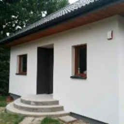 Biały, otynkowany budynek z brązowymi oknami i drzwiami w Krakowie, otoczony zielenią. Widoczne schody wejściowe wyłożone kamieniem.