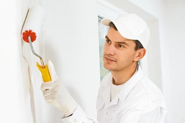 Malarz w białym uniformie i czapce maluje ścianę wałkiem z czerwonymi elementami mocującymi, patrząc w prawo, w jasnym pomieszczeniu.