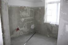 Surowe wnętrze łazienki w trakcie remontu: szare, nierówne ściany z widocznymi śladami po instalacjach, rurka na podłodze, okno z białymi firankami w tle.