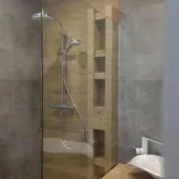 Nowoczesna kabina prysznicowa z drewnopodobnymi płytkami i wbudowanymi półkami, widoczna umywalka nablatowa z białą armaturą.