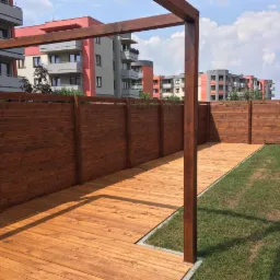 Drewniany taras z pergolą i ogrodzeniem z desek, widok na nowoczesne budynki mieszkalne w tle.