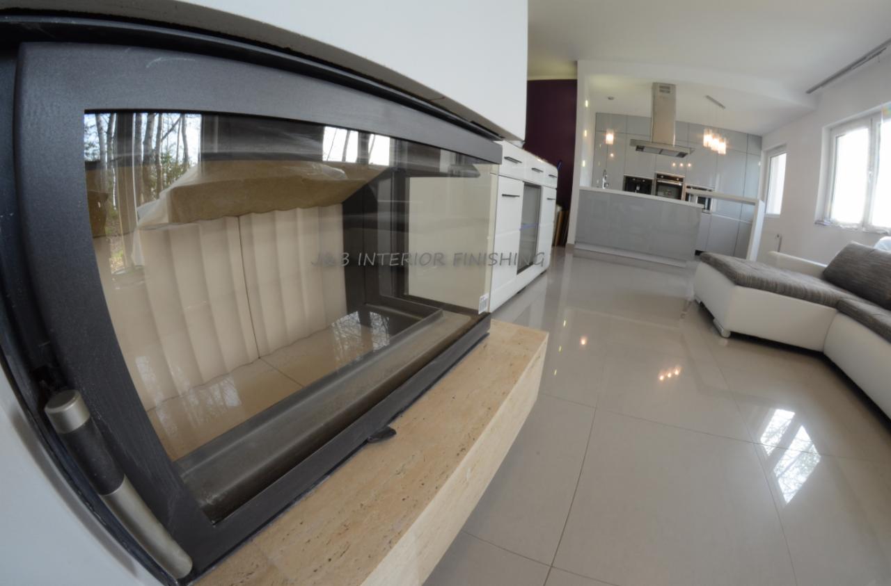 Nowoczesny wkład kominkowy z czarną ramą i jasnym wykończeniem paleniska, widok na salon z błyszczącą podłogą, białą zabudową meblową i szarą kuchnią w tle.