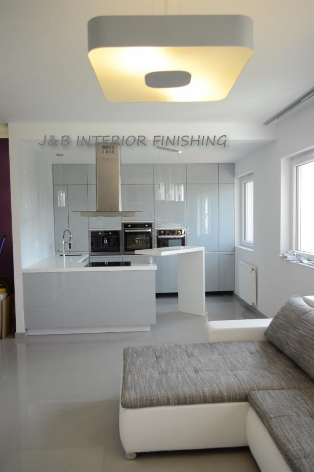 Jasnoszara kuchnia z wyspą, nowoczesne oświetlenie sufitowe, widoczny fragment szarej sofy w salonie.