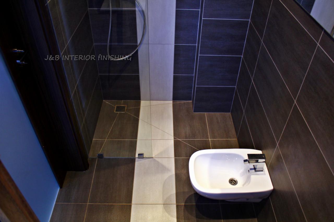 Nowoczesna łazienka z ciemnymi płytkami, białą armaturą i szklaną kabiną prysznicową; widoczny logo firmy 'J&B Interior Finishing' na ścianie.