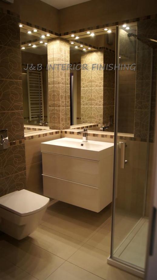 Wykończona łazienka w odcieniach beżu i brązu z podwieszaną szafką umywalkową, toaletą i kabiną prysznicową, oświetlona punktowymi lampami w suficie.