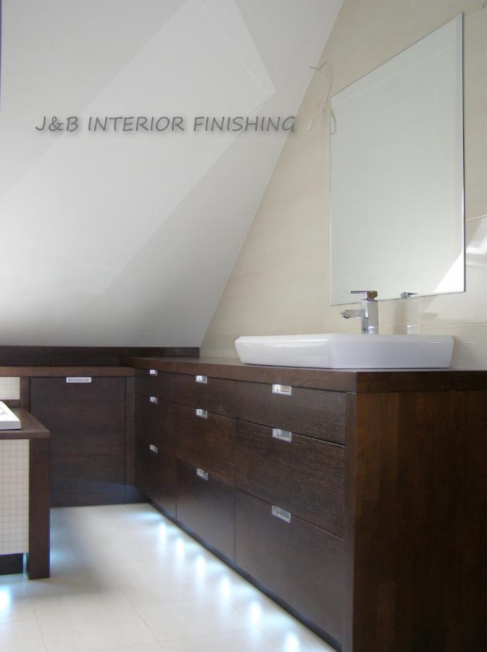 Nowoczesna łazienka na poddaszu z ciemnymi drewnianymi szafkami pod umywalką, oświetleniem LED u dołu i prostokątnym lustrem.