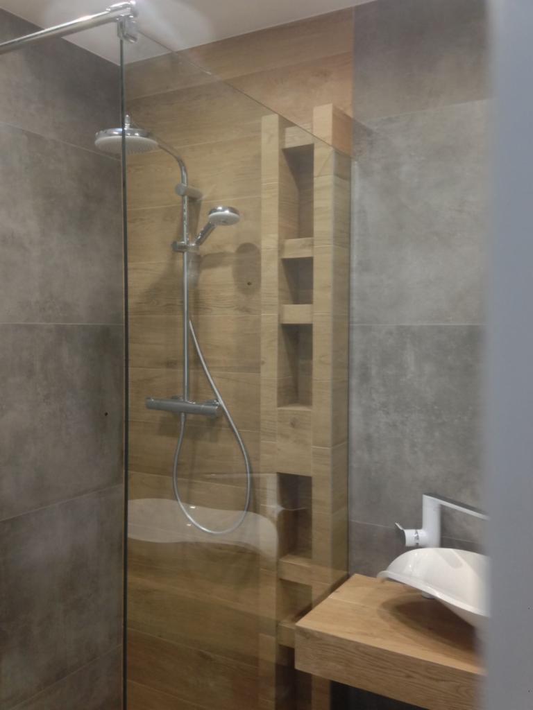 Nowoczesna kabina prysznicowa z drewnopodobnymi płytkami i wbudowanymi półkami, widoczna umywalka nablatowa z białą armaturą.