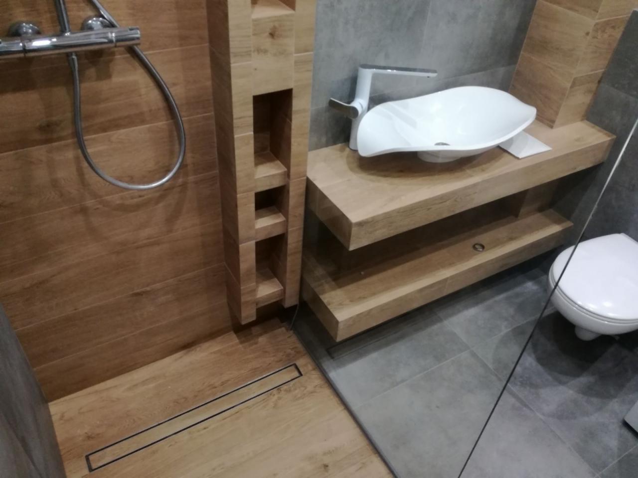 Nowoczesna łazienka z drewnopodobnymi płytkami na ścianach i podłodze, białą umywalką w kształcie liścia na drewnianym blacie i szarą podłogą z płytek.