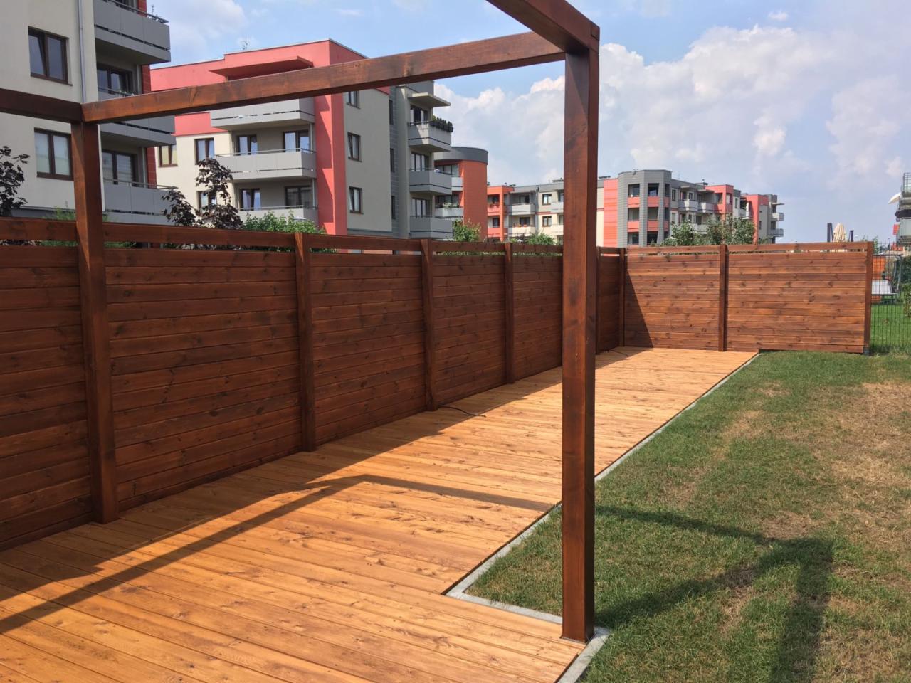 Drewniany taras z pergolą i ogrodzeniem z desek, widok na nowoczesne budynki mieszkalne w tle.