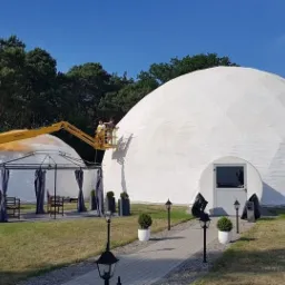 Pracownicy na podnośniku koszowym aplikują białą powłokę na kopułę geodezyjną, widoczna tekstura materiału, w tle druga kopuła i altana.
