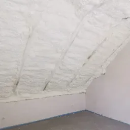 Poddasze po ociepleniu pianką PUR, widoczne krokwie pokryte grubą warstwą białej piany, ściany pomalowane na jasny kolor, zabezpieczone niebieską taśmą malarską przy podłodze.