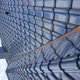 Błyszczące, ciemne dachówki ceramiczne ułożone na dachu domu, widoczne z dołu pod kątem, z fragmentem lukarny i niebem w tle.