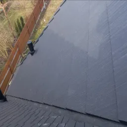 Czarny dach z łupka z dwoma kominkami wentylacyjnymi, widok z góry na tle płotu i drzew bez liści.