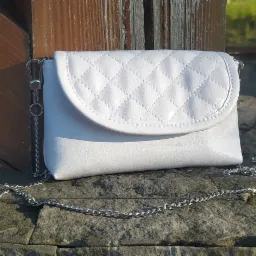 Biała, pikowana torebka z łańcuszkiem, leżąca na kamiennym murku w plenerze, z drewnianym elementem w tle.