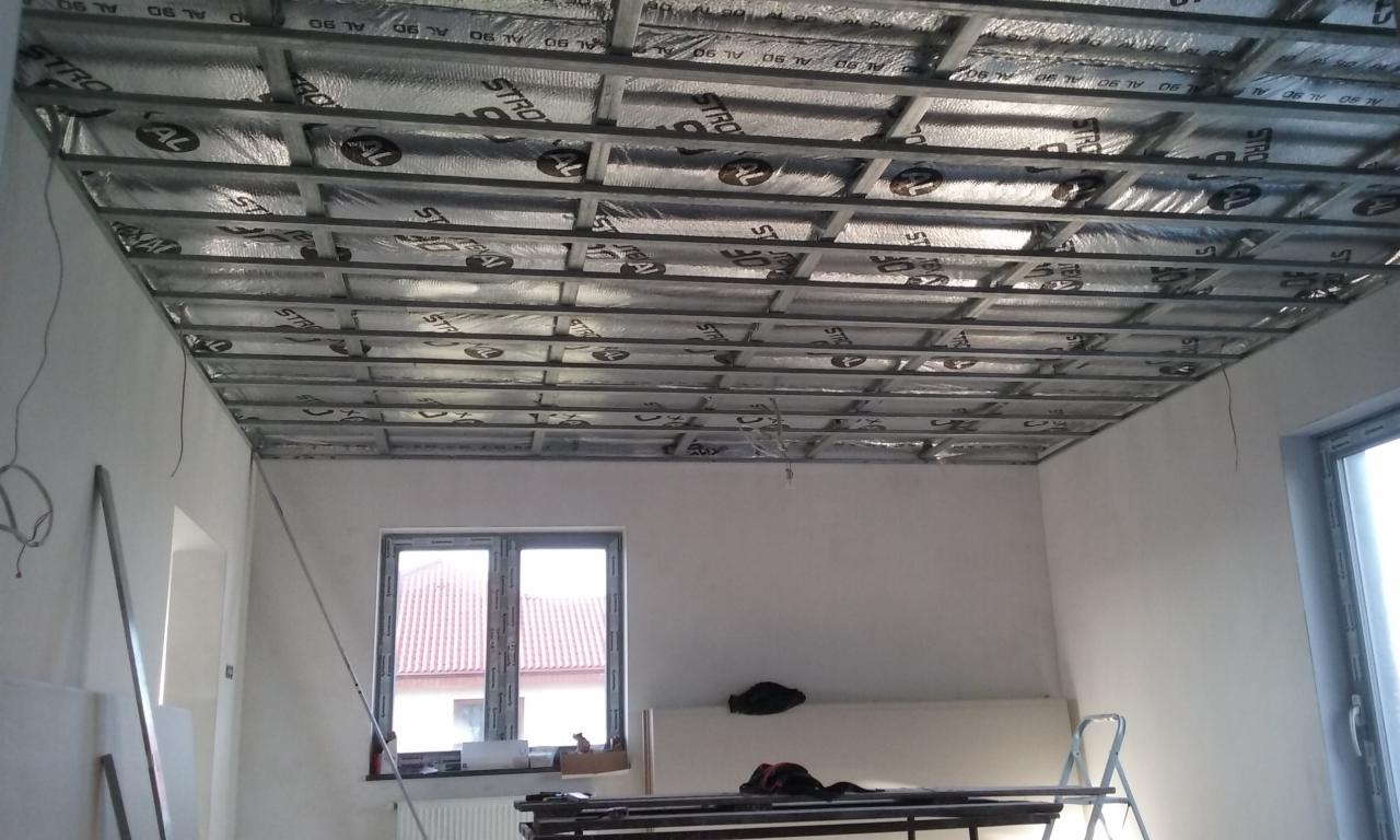 Montaż metalowej konstrukcji sufitu podwieszanego z izolacją termiczną w jasnym pomieszczeniu z oknami, widoczne elementy instalacji elektrycznej i drabina.