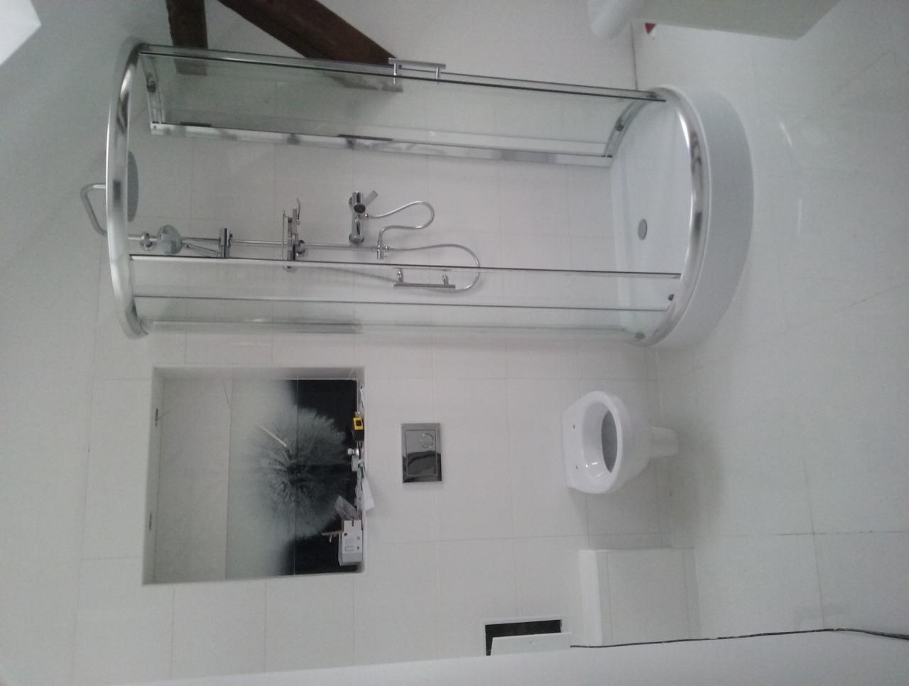 Biała łazienka w trakcie remontu: kabina prysznicowa z zaokrąglonymi drzwiami, podwieszana toaleta i otwór na ścianie z widoczną grafiką dmuchawca oraz wystającymi przewodami elektrycznymi.