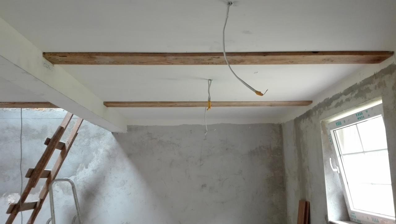 Surowy stan pokoju z drewnianymi belkami pod sufitem, widoczne przewody elektryczne i drabina oparta o ścianę, ściany w szarym tynku, okno z PCV.