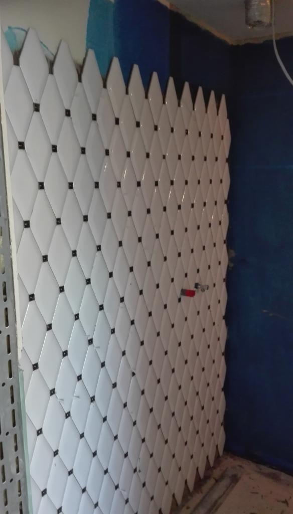 Ściana wyłożona białymi, romboidalnymi płytkami z czarnymi kwadratowymi wstawkami, tworząca dekoracyjny wzór z ząbkowanym wykończeniem u góry, w tle fragment niebieskiej ściany.