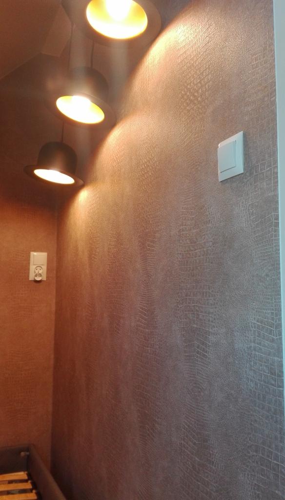 Ściana z fakturą skóry krokodyla w odcieniach brązu oświetlona trzema okrągłymi lampami sufitowymi, widoczny włącznik światła i gniazdko elektryczne.
