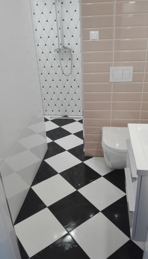Nowoczesna łazienka z czarno-białą szachownicą na podłodze, kabina prysznicowa z białymi płytkami zdobionymi czarnymi kwadratami, beżowe płytki na ścianie, biała toaleta i umywalka.