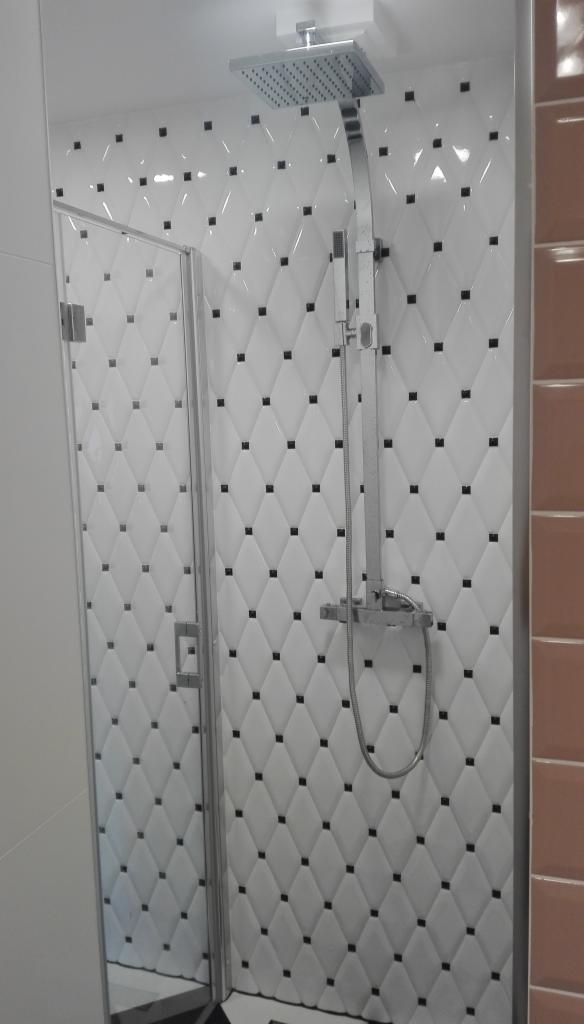 Wykończona kabina prysznicowa z białymi, wypukłymi płytkami w romby z czarnymi akcentami, chromowana armatura i szklane drzwi.