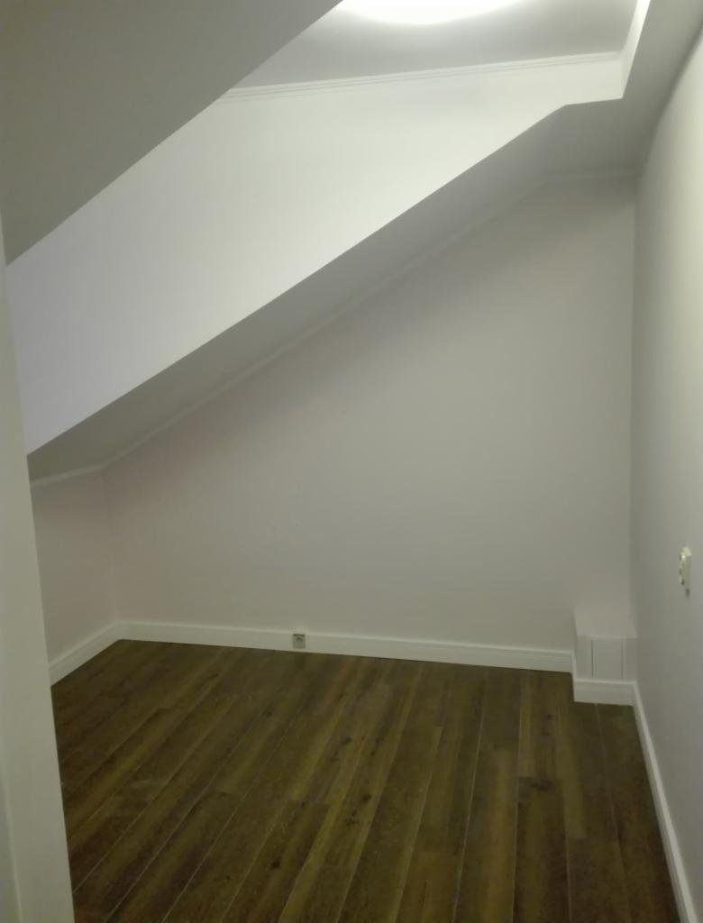 Poddasze z drewnianą podłogą w odcieniach brązu i białymi ścianami, widoczne skosy i listwy przypodłogowe, oświetlenie sufitowe.