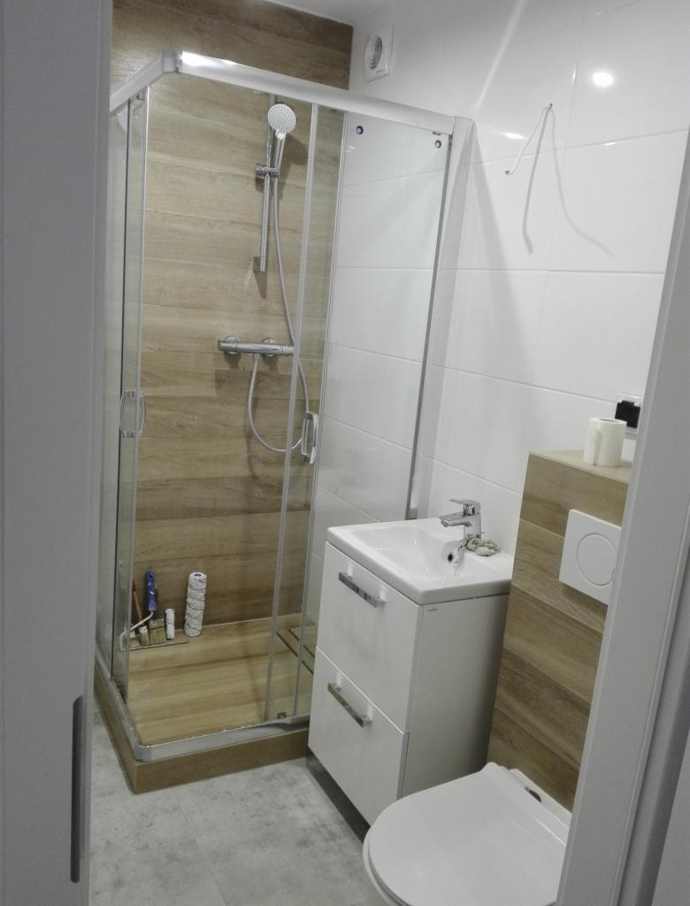 Nowoczesna łazienka z kabiną prysznicową z drewnopodobnymi płytkami, białą umywalką z szafką i wiszącą toaletą. Na półce w kabinie prysznicowej widoczne akcesoria łazienkowe.