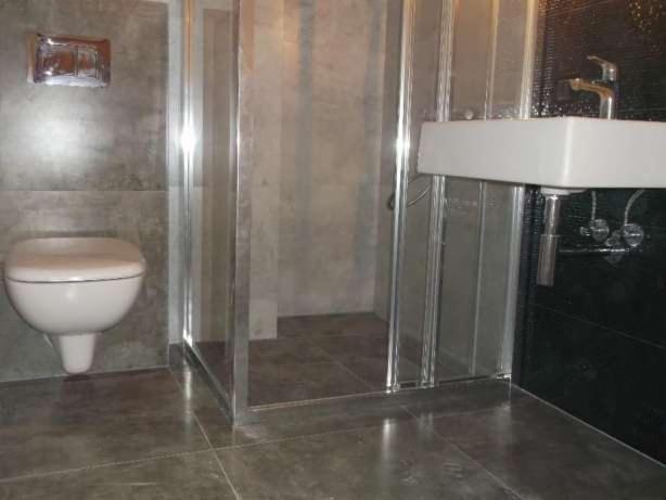 Szara łazienka z kabiną prysznicową, podwieszaną toaletą i umywalką nablatową na czarnej ścianie, widoczne rury odpływowe.