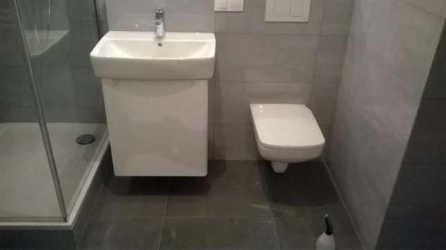 Nowoczesna łazienka z szarymi płytkami, wiszącą umywalką z szafką, podwieszanym WC i kabiną prysznicową z przezroczystego szkła.