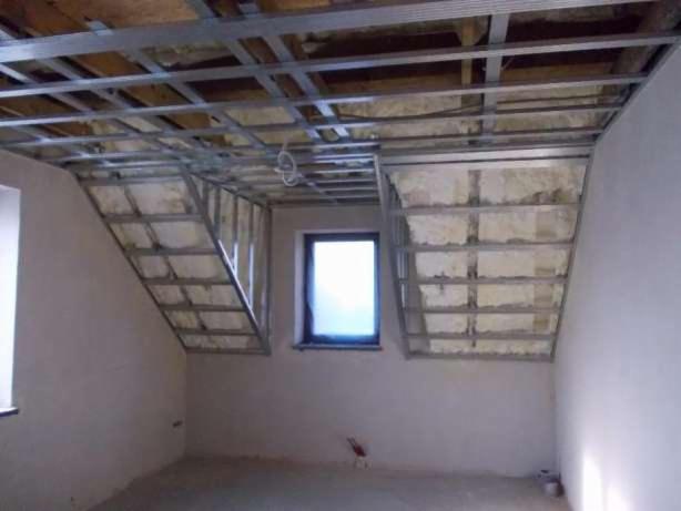 Montaż metalowej konstrukcji sufitu podwieszanego na poddaszu ze skośnymi ścianami, widoczna izolacja z wełny mineralnej, okno w ścianie szczytowej, surowy stan wykończenia.