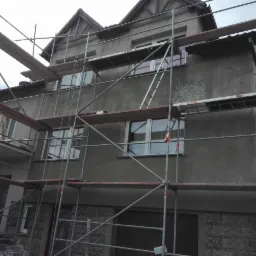 Dom w trakcie tynkowania elewacji, widoczne rusztowania i fragmenty surowego tynku na ścianie.