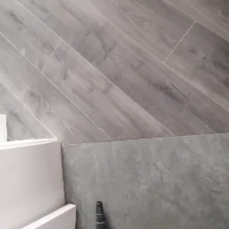 Szare panele podłogowe ułożone skośnie, stykające się z szarymi płytkami ściennymi, fragment białej zabudowy meblowej w lewym dolnym rogu.