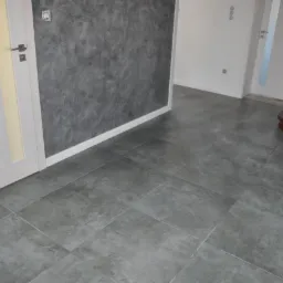 Szare, kwadratowe płytki podłogowe ułożone w korytarzu z szarą ścianą o fakturze betonu i białymi drzwiami z przeszkleniami, widoczne schody drewniane.