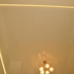 Ambasada Remontowa - Błyszczący, beżowy sufit z wbudowanym oświetleniem LED wzdłuż krawędzi, w centrum widoczny ozdobny, kryształowy żyrandol.