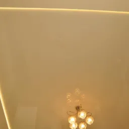 Błyszczący, beżowy sufit z wbudowanym oświetleniem LED wzdłuż krawędzi, w centrum widoczny ozdobny, kryształowy żyrandol.