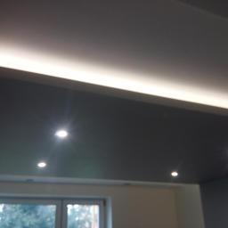 Ambasada Remontowa - Podwieszany sufit z oświetleniem LED i punktowymi lampami halogenowymi, widok z dołu na tle okna.