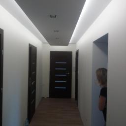 Ambasada Remontowa - Korytarz po remoncie z drewnopodobną podłogą, białymi ścianami, ciemnymi drzwiami z poziomymi przeszkleniami i podwieszanym sufitem z oświetleniem LED, widok z perspektywy osoby stojącej w progu...