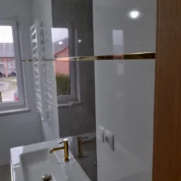 Nowoczesna łazienka w trakcie wykańczania: biała umywalka z baterią w kolorze złota, fragment grzejnika drabinkowego, szare płytki imitujące marmur na ścianie, widoczne kable wystające ze ściany.