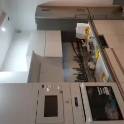 Nowoczesna, biała kuchnia z zabudowanymi sprzętami, okapem i panelem ściennym z motywem panoramy miasta. Widoczne oświetlenie punktowe na suficie i fragment lodówki.