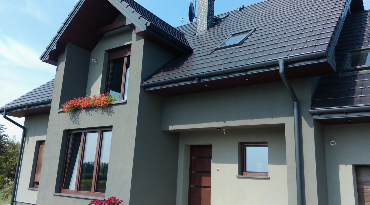 Dwukondygnacyjny dom z szarą elewacją, ciemnym dachem, brązowymi oknami i balkonem z czerwonymi kwiatami, widziany z zewnątrz w słoneczny dzień.