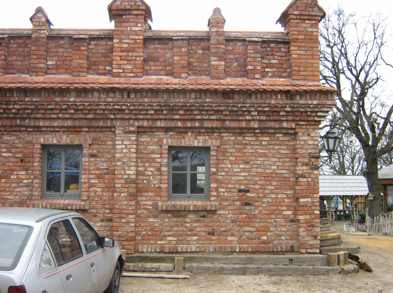 Murowana ściana budynku z czerwonej cegły z dwoma szarymi oknami, dekoracyjnymi elementami na górze i fragmentem srebrnego samochodu w lewym dolnym rogu.