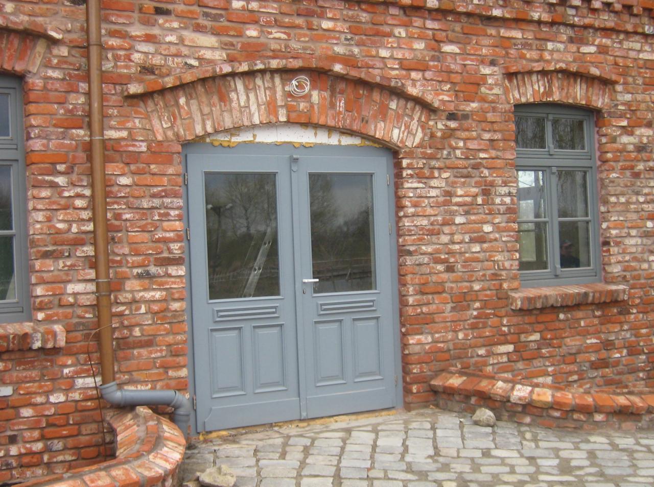 Murowany budynek z cegły z szarymi drzwiami i oknami, widoczny fragment rynny i kamienna kostka brukowa przed wejściem.