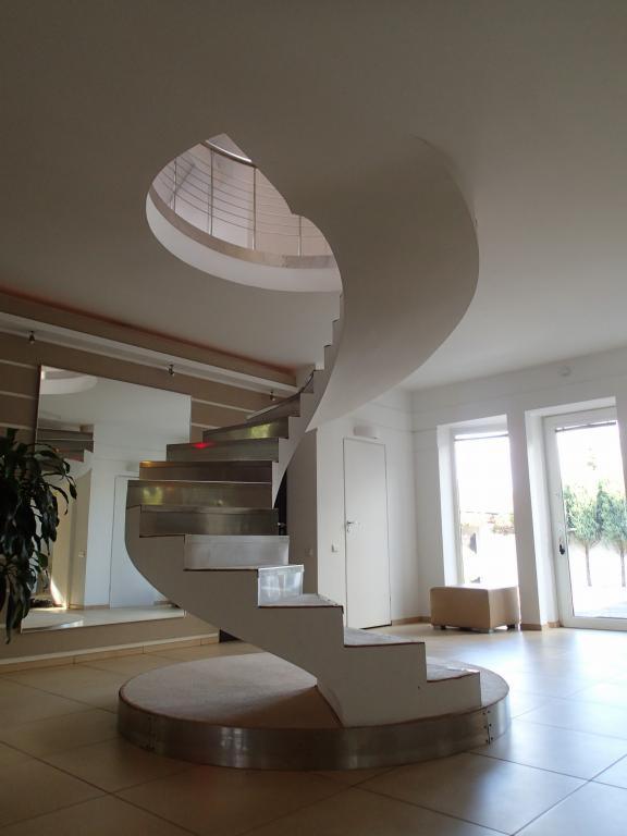 Spiralne schody z metalowymi stopniami w nowoczesnym wnętrzu z dużymi oknami i lustrem.