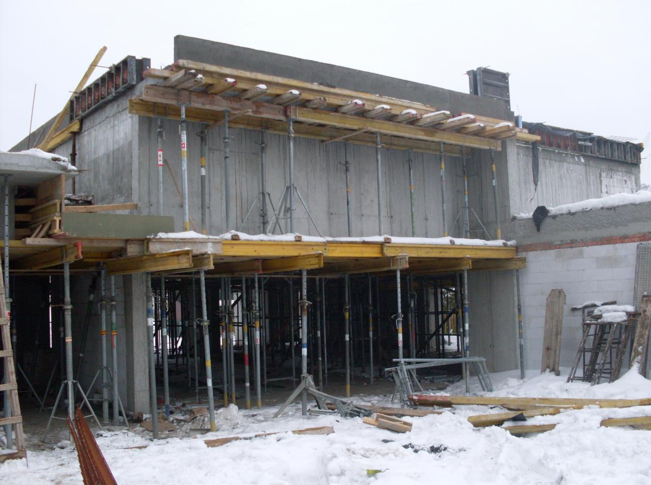 Surowy stan budynku w trakcie budowy, betonowe ściany z drewnianym szalunkiem wsparte metalowymi stemplami, zasypane śniegiem.