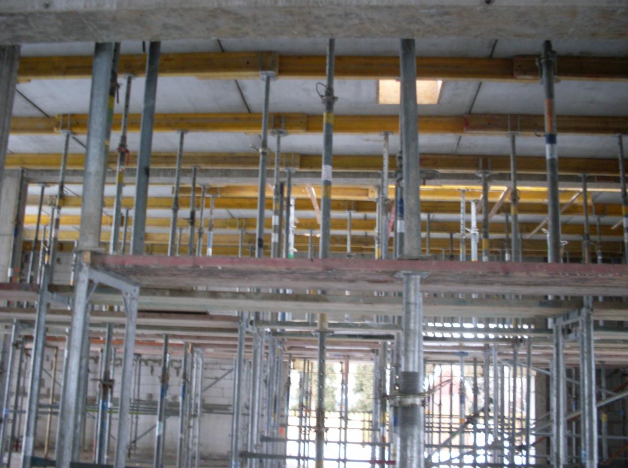 Szare, metalowe stemple podtrzymujące żółte, drewniane szalunki stropu w trakcie budowy domu murowanego, widoczne z poziomu rusztowania.