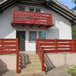 Dom z drewnianą balustradą balkonu i ogrodzeniem tarasu z surowego drewna w kolorze wiśniowym, schody wejściowe wyłożone płytkami.