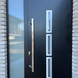 Czarne drzwi wejściowe z długim, pionowym uchwytem ze stali nierdzewnej, trzema wąskimi, prostokątnymi przeszkleniami i bocznym panelem szklanym, osadzone w murze z jasnej cegły.