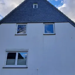 Elewacja domu z białym tynkiem, trzema oknami, w tym jednym z plisami, oraz dachem pokrytym szarym łupkiem.